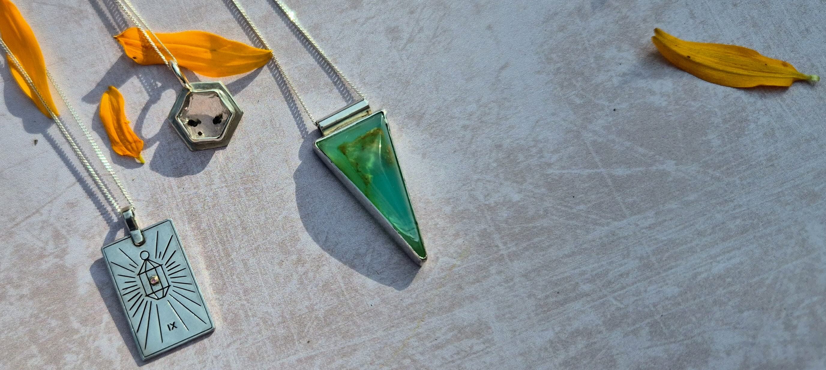 Three distinct silver-toned pendant necklaces arranged on a textured light gray surface with yellow flower petals. The necklaces feature a silver rectangular pendant with an engraved sunburst and symbol, a silver hexagonal pendant, and a silver triangular pendant showcasing a green and gold marbled stone.